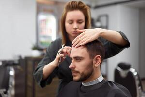 stilig blå ögon man Sammanträde i barberare affär. hårstylist frisör kvinna skärande hans hår. kvinna barberare. foto