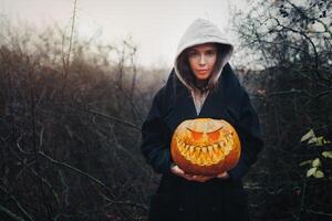 Lycklig halloween porträtt av svart modern livsstil häxa i huva och svart täcka mantel på trä- buske skog bakgrund. innehav pumpa i händer foto