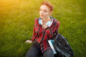 ung skön studerande bär en rutig skjorta hörlurar och en ryggsäck vilar på de gata mellan klasser foto
