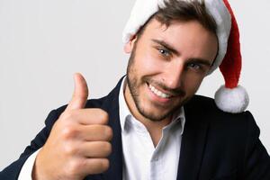 ung stilig caucasian kille i företag kostym och santa hattar står på vit bakgrund i studio smilie och som visar tummen upp. foto