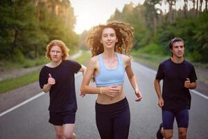 en grupp av tre människor idrottare ett flicka och två män springa på ett asfalt väg i en tall skog. foto