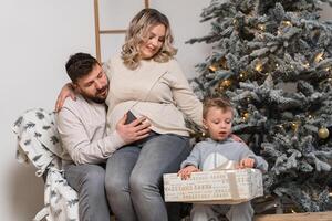 jul familj lycka porträtt av pappa, gravid mamma och liten son Sammanträde fåtölj på Hem nära jul träd kram leende foto