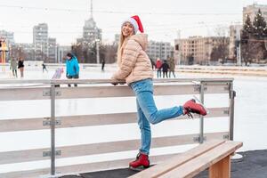 skön härlig medelålders flicka blond hår värma vinter- jackor stickat handske står is rink bakgrund stad fyrkant. jul humör livsstil foto