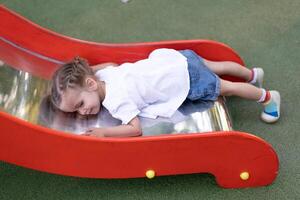 liten caucasian flicka lögner på en glida på de lekplats trött av en ledsen sommar dag. foto