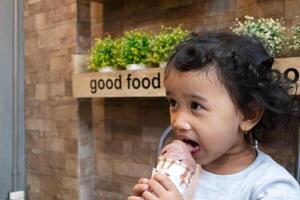 ett asiatisk liten flicka med lockigt hår äter en choklad is grädde i de Kafé foto