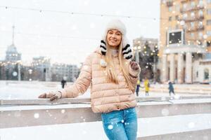 skön härlig medelålders flicka blond hår värma vinter- jackor stickat handske står is rink bakgrund stad fyrkant. jul humör livsstil foto