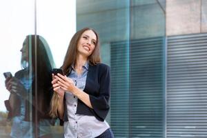 företag kvinna 35 år klädd rand skjorta och svart jacka med lång hår stående nära kontor byggnad utomhus- använda sig av smartphone foto