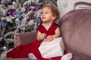 en skön söt liten flicka klädd i ett elegant kväll röd klänning sitter på de soffa och öppnas en ny år gåva. med jul träd på de bakgrund foto