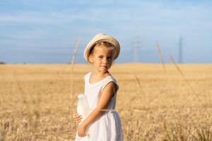 liten flicka stående vete fält innehav flaska av mjölk i hand klädd vit klänning och sugrör hatt foto