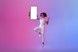 energisk svart kvinna Hoppar med tom smartphone skärm på neon bakgrund foto
