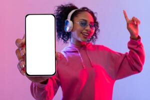Lycklig kvinna med hörlurar, visa upp smartphone och dans, färgrik bakgrund foto