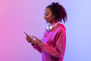 leende svart lady använder sig av smartphone med hörlurar, rosa luvtröja foto