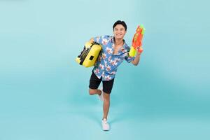 Lycklig stilig asiatisk turist man i sommar klädespersedlar med vatten pistol och resväska i blå Färg studio isolerat bakgrund foto