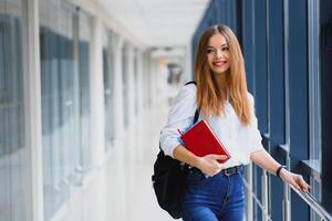 positivitet skön flicka leende på kamera, stående på korridor med anteckningar som ryggsäck, gående till lektion. Lycklig brunett kvinna studerande studerar i lyx universitet. foto