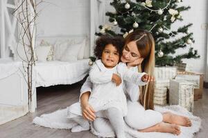 glad jul och Lycklig högtider glad mamma och henne söt dotter på jul träd. förälder och liten barn har roligt nära jul träd inomhus. kärleksfull familj med presenterar i rum. foto