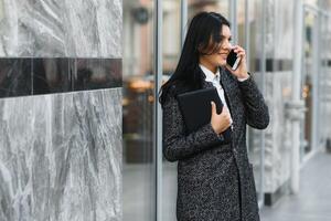 attraktiv ung kvinna i de stad. företag lady är leende medan talande på en smart telefon i främre av modern företag Centrum foto