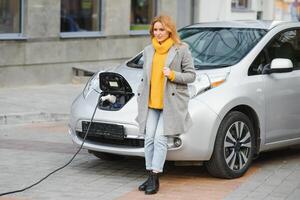 kvinna med telefon nära ett uthyrning elektrisk bil. fordon laddad på de laddning station foto