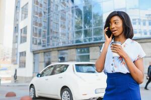 skön afrikansk amerikan flicka stående på gata med mobiltelefon och kaffe i händer medan lyckligt ser åt sidan. porträtt av afrikansk amerikan lady på gata. foto