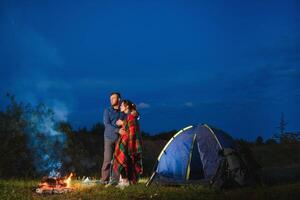 kärleksfull par vandrare njuter varje Övrig, stående förbi lägereld på natt under kväll himmel nära träd och tält. romantisk camping nära skog i de bergen foto