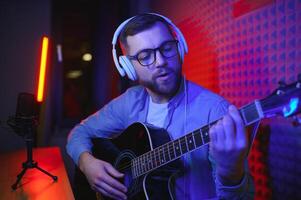 en ung eleganta kille med en skägg bär tillfällig eller musik lärare spelar gitarr i främre av smartphone kamera. uppkopplad gitarr Träning eller sång- lektion foto