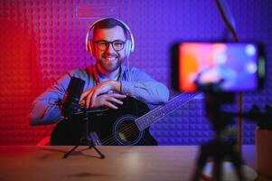 en ung eleganta kille med en skägg bär tillfällig eller musik lärare spelar gitarr i främre av smartphone kamera. uppkopplad gitarr Träning eller sång- lektion foto