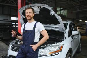 skäggig mekaniker i overall stående i garage av en bil salong och innehav läsplatta. han är handla om till diagnostisk bryta ner. foto