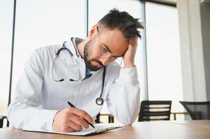 stilig läkare man bär medicinsk enhetlig Sammanträde på hans arbetsplats trött innehav hans huvud känsla Trötthet och huvudvärk. påfrestning och frustration begrepp foto