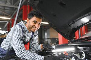 indisk bil mekaniker stående och arbetssätt i service station. bil specialister granskning de lyft bil. professionell reparatörer bär mekaniker enhetlig i blå Färg. foto