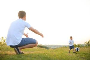 far med en liten son spelar fotboll på de grön gräs i de parkera. Lycklig familj har roligt och spelar fotboll på en grön gräs- gräsmatta på en solig dag. familj begrepp, fars dag foto