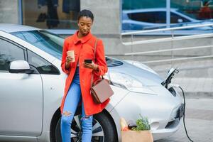 använder sig av smartphone medan väntar. afrikansk amerikan flicka på de elektrisk bilar avgift station på dagtid. varumärke ny fordon foto