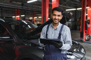 indisk bil mekaniker stående och arbetssätt i service station. bil specialister granskning de lyft bil. professionell reparatörer bär mekaniker enhetlig i blå Färg. foto