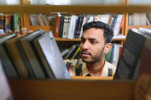 porträtt av glad manlig internationell indisk studerande med ryggsäck, inlärning Tillbehör stående nära bokhyllor på universitet bibliotek eller bok Lagra under ha sönder mellan lektioner. utbildning begrepp foto