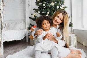 glad jul och Lycklig högtider glad mamma och henne söt dotter på jul träd. förälder och liten barn har roligt nära jul träd inomhus. kärleksfull familj med presenterar i rum. foto