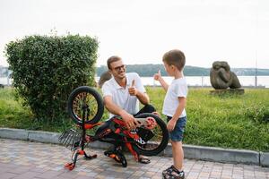 Lycklig far och hans son har roligt tillsammans på de grön parkera, fixering cykel tillsammans. fars dag. foto