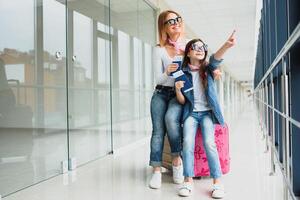 Lycklig och eleganta mamma med henne dotter på de flygplats ser ut av flygplats terminal fönster till ser plan och Övrig. foto