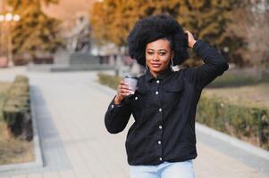 porträtt av en leende ung afrikansk amerikan flicka med pigtails med kaffe gående i de gata på en solig dag. utomhus- Foto. foto