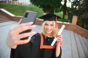 Lycklig kvinna studerande innehav universitet diplom medan tar selfie efter gradering ceremoni. foto