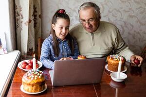 farfar och barnbarn är talande via video länk till deras vänner. dekorerad tabell med färgrik ägg och kaka. chattar under de covid pandemi och de påsk högtider foto