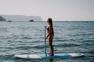 kvinna hav supera. stänga upp porträtt av Lycklig ung caucasian kvinna med lång hår ser på kamera och leende. söt kvinna porträtt i en blå bikini Framställ på supera styrelse i de hav foto