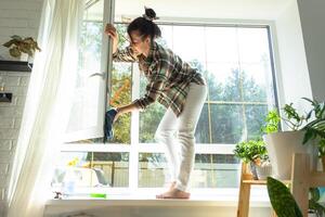 kvinna manuellt tvättar de fönster av de hus med en trasa med spray rengöringsmedel och mopp inuti de interiör med vit gardiner. återställa beställa och renlighet i de vår, rengöring serva foto