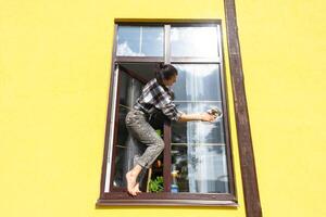 en kvinna manuellt tvättar de fönster av de hus med en trasa med en spray rengöringsmedel och en mopp utanför. säkerhet på höjd, återställa beställa och renlighet i de vår, rengöring service foto