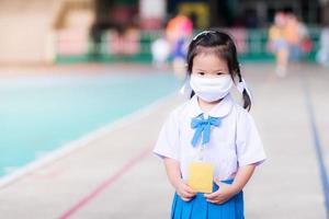 barn som bär vit tyg ansiktsmask i skoluniform står. barn går i skolan med ett nytt sätt att leva nytt normalt. söt unge tittar på kameran. dagis småbarn ålder 3 år. foto