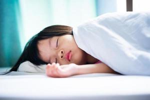 asiatiska barn sover på en vit mjuk säng. kid flicka täcker sin kropp med en vit filt. bebis i åldern 4-5 år. foto