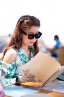 porträtt av asiatiska vackra kvinnor som sitter i restaurangen vid stranden. tonåring kvinna håller bok och letar matmeny. person som bär solglasögon. sommartid. foto