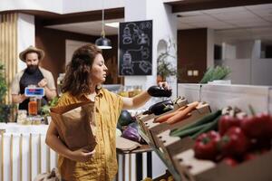 kvinna i noll avfall affär köp av organisk lokalt vuxen grönsaker, plockning mogen äggplantor. kund i lokal- matvaror affär ser till köpa friska mat, använder sig av papper väska till undvika enda använda sig av plast foto