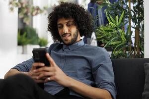 företag projekt chef skrivning meddelande till medarbetare använder sig av smartphone i företag kontor. Start upp ung arab entreprenör avkopplande och chattar i social media på smartphone foto