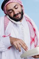 muslim man bär traditionell slöja uppringning siffra på fast telefon telefon. arab person klädd i kulturell islamic kläder innehav retro telefon och framställning ring upp närbild foto