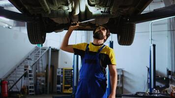 noggrann serviceman arbetssätt på suspenderad bil i garage, kontroll komponenter under rutin- underhåll. man i bil reparera affär gående under fordon, undersökande den använder sig av arbete ljus foto