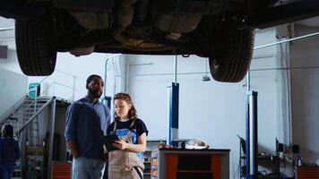 kvalificerad ingenjör i reparera affär som visar kund Vad behov till vara ändrats på hans bil. garage anställd inspekterande fordon placerad på över huvudet hiss, talande med afrikansk amerikan klient foto