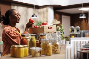 Foto fokus på de leende svart kvinna i en bio-mat Lagra granskning ett organisk gårdsodlad tomat. klient av afrikansk amerikan etnicitet glatt beundrar färsk producera i de eco vänlig marknadsföra.